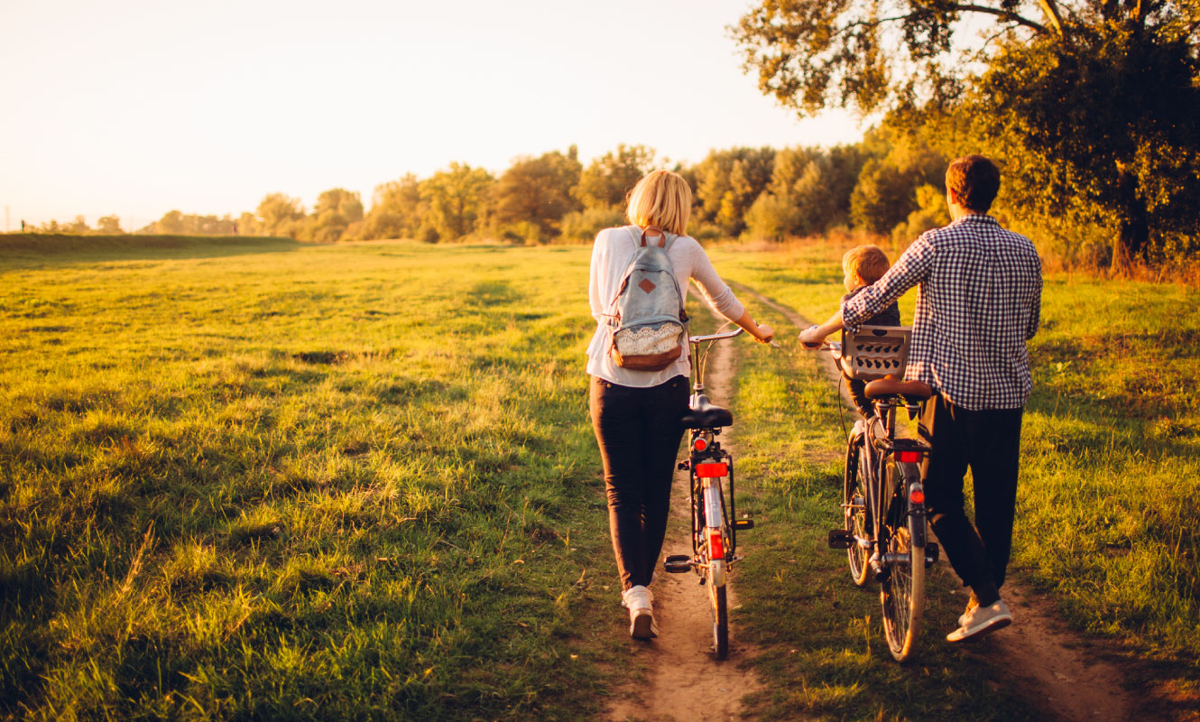 A young couple and their baby have their backs towards the camera. They are pushing bicycles on a dirt road in a green field and the sun is setting. 